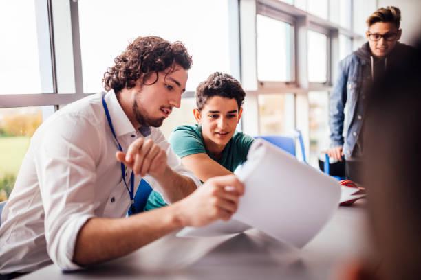 Teacher showing a student some results on his paperwork. The student looks happy as they sit side by side at a table. Another student looks on.